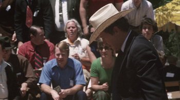 Movie still from “Cockfighter” (1974), directed by Monte Hellman – A man in a cowboy hat is standing in front of a group of people; Medium shot, Over the shoulder angle