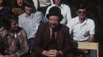 Movie still from “Cockfighter” (1974), directed by Monte Hellman – A man in a suit and hat sitting in front of a group of people; Medium shot, Over the shoulder angle