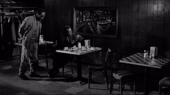 Movie still from “Coffee and Cigarettes” (2003), directed by Jim Jarmusch – A man sitting at a table in front of a painting; Wide shot, High angle