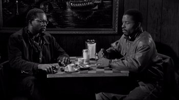 Movie still from “Coffee and Cigarettes” (2003), directed by Jim Jarmusch – Two men are sitting at a table in front of a painting; Medium shot, Over the shoulder angle