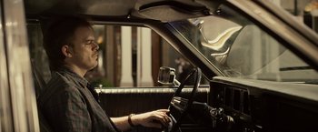 Movie still from “Cold in July” (2014), directed by Jim Mickle – A man sitting in the driver's seat of a car; Medium shot, Over the shoulder angle