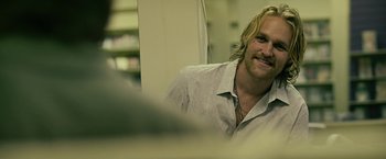 Movie still from “Cold in July” (2014), directed by Jim Mickle – A man with long blonde hair smiling for the camera; Close Up shot, Over the shoulder angle