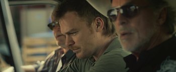 Movie still from “Cold in July” (2014), directed by Jim Mickle – A man sitting next to two other men in a car; Close Up shot, Over the shoulder angle