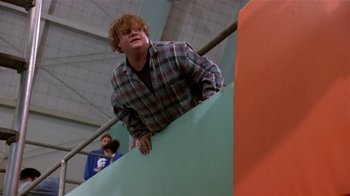 Movie still from “Coneheads” (1993), directed by Steve Barron – A man leaning up against a railing in an indoor arena; Medium shot, Low angle