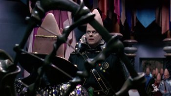 Movie still from “Coneheads” (1993), directed by Steve Barron – A man in a black jacket is sitting in front of a bunch of clothes; Medium shot, Over the shoulder angle