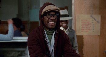 Movie still from “Cooley High” (1975), directed by Michael Schultz – A man wearing a hat and glasses smiling for the camera; Medium shot, Over the shoulder angle