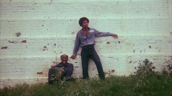 Movie still from “Coonskin” (1974), directed by Ralph Bakshi – Two men standing next to each other in front of a brick wall; Wide shot, Low angle