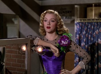Movie still from “Cover Girl” (1944), directed by Charles Vidor – A woman in a purple dress holding a purple flower; Medium shot, Low angle