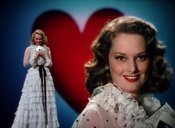 Movie still from “Cover Girl” (1944), directed by Charles Vidor – A woman in a white dress standing in front of a red heart; Medium shot, Low angle