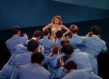 Movie still from “Cover Girl” (1944), directed by Charles Vidor – A group of people sitting around a woman in a crowd; Wide shot, High angle
