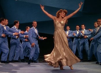 Movie still from “Cover Girl” (1944), directed by Charles Vidor – A woman in a gold dress dancing with a group of men in blue suits; Wide shot, Low angle