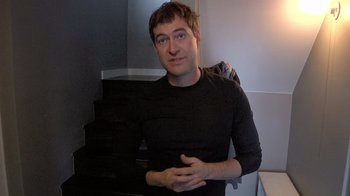 Movie still from “Creep” (2014), directed by Patrick Brice – A man in a black shirt is sitting on steps; Medium shot, Low angle