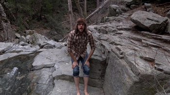 Movie still from “Creep” (2014), directed by Patrick Brice – A man in a plaid shirt is squatting on some rocks; Wide shot, Overhead angle