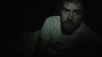 Movie still from “Creep” (2014), directed by Patrick Brice – A man with a goatee and a white t - shirt is laying in a bed; Close Up shot, High angle