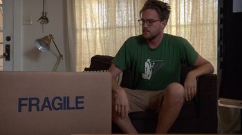 Movie still from “Creep” (2014), directed by Patrick Brice – A man sitting on a couch next to an open box; Medium shot, Low angle