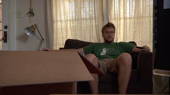 Movie still from “Creep” (2014), directed by Patrick Brice – A man sitting on top of a brown couch next to boxes; Medium shot, High angle