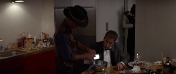 A man in a cowboy hat is lighting a candle on a man in a tuxedo