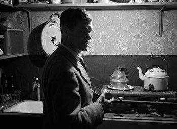Movie still from “Crossfire” (1947), directed by Edward Dmytryk – A black and white photo of a man smoking a cigarette in a kitchen; Medium shot, Low angle