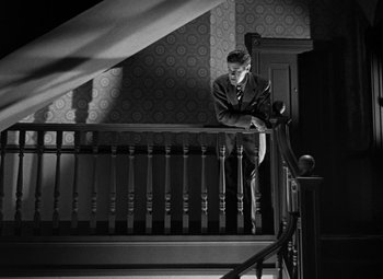 Movie still from “Crossfire” (1947), directed by Edward Dmytryk – A black and white photo of a man reading a newspaper on the stairs; Wide shot, Low angle
