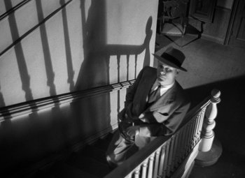 Movie still from “Crossfire” (1947), directed by Edward Dmytryk – A black and white photo of a man sitting on a stair case railing; Wide shot, Low angle
