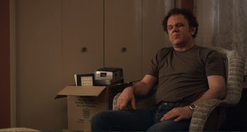 Movie still from “Cyrus” (2010), directed by Jay Duplass – A man sitting on a couch next to a box; Medium shot, Low angle