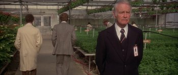 Movie still from “Damien: Omen II” (1978), directed by Mike Hodges – A man wearing a suit and tie in a greenhouse; Medium shot, Over the shoulder angle
