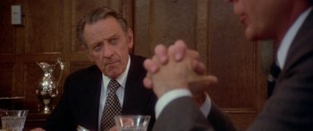 Movie still from “Damien: Omen II” (1978), directed by Mike Hodges – A man sitting at a table with a glass of water and another man clasping his hands; Close Up shot, Over the shoulder angle