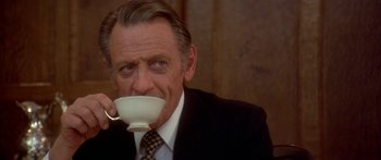 Movie still from “Damien: Omen II” (1978), directed by Mike Hodges – An older man wearing a suit and tie drinking from a white cup; Close Up shot, Low angle