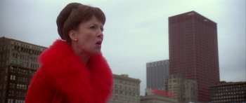 Movie still from “Damien: Omen II” (1978), directed by Mike Hodges – A woman wearing a red fur stole standing in front of some buildings; Close Up shot, Low angle