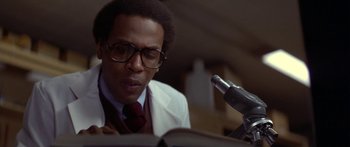 Movie still from “Damien: Omen II” (1978), directed by Mike Hodges – A man in a white lab coat reading a book in front of a microphone; Close Up shot, Low angle