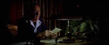 Movie still from “Damien: Omen II” (1978), directed by Mike Hodges – An older man sitting at a table with papers in front of him; Medium shot, Over the shoulder angle