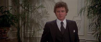 Movie still from “Damien: Omen II” (1978), directed by Mike Hodges – A man in a suit and tie standing in front of a plant; Medium shot, Low angle