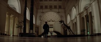 Movie still from “Damien: Omen II” (1978), directed by Mike Hodges – A man in a suit walking in a building with a dinosaur statue; Extreme Wide shot, Low angle