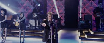 Movie still from “Danny Collins” (2015), directed by Dan Fogelman – A man in a black suit and tie holding a microphone; Wide shot, High angle