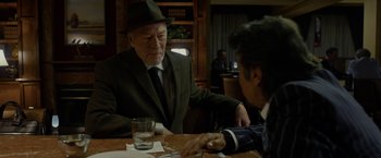 Movie still from “Danny Collins” (2015), directed by Dan Fogelman – Two men sitting at a wooden table in front of a glass of water; Medium shot, Over the shoulder angle