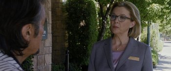 Movie still from “Danny Collins” (2015), directed by Dan Fogelman – A woman in a gray suit and glasses standing in front of a brick wall; Close Up shot, Over the shoulder angle