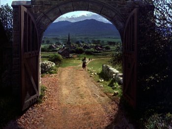 Movie still from “Darby O'Gill and the Little People” (1959), directed by Robert Stevenson – A person riding a motorcycle on a dirt road under an arched gate; Extreme Wide shot, High angle