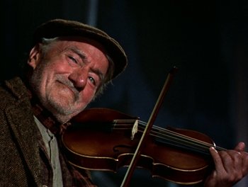 Movie still from “Darby O'Gill and the Little People” (1959), directed by Robert Stevenson – An older man holding a violin in front of a dark background; Close Up shot, Low angle