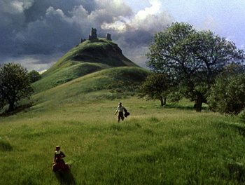 Movie still from “Darby O'Gill and the Little People” (1959), directed by Robert Stevenson – Two people walking in a grassy field with a castle in the background; Extreme Wide shot, High angle