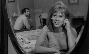 Movie still from “Darling” (1965), directed by John Schlesinger – A black and white photo of a woman sitting on a bed with a man sitting in the background; Close Up shot, Over the shoulder angle