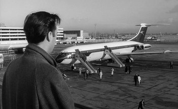 Movie still from “Darling” (1965), directed by John Schlesinger – A black and white photo of a man looking at an airplane on the tarmac; Wide shot, Low angle
