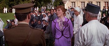 Movie still from “Darling Lili” (1970), directed by Blake Edwards – A woman in a purple dress standing in front of a crowd; Medium shot, Over the shoulder angle