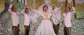Movie still from “Darling Lili” (1970), directed by Blake Edwards – A woman in a white dress and hat singing in front of a group of men; Medium shot, Low angle