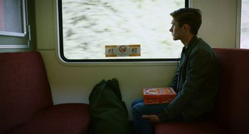 Movie still from “Dating Amber” (2020), directed by David Freyne – A man sitting on a train holding a box of pizza; Medium shot, Over the shoulder angle