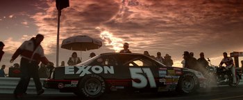 Movie still from “Days of Thunder” (1990), directed by Tony Scott – A group of people standing around a race car at sunset; Extreme Wide shot, Low angle