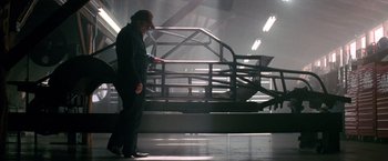 Movie still from “Days of Thunder” (1990), directed by Tony Scott – A man standing next to a car in a building; Wide shot, Low angle