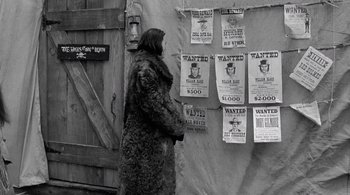 Movie still from “Dead Man” (1995), directed by Jim Jarmusch – A woman in a fur coat standing next to a wall of wanted posters; Wide shot, High angle