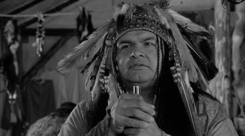 Movie still from “Dead Man” (1995), directed by Jim Jarmusch – A man in a native american headdress holding a pipe; Close Up shot, Over the shoulder angle