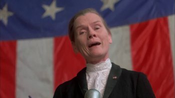 Movie still from “Death Race 2000” (1975), directed by Paul Bartel – A man in a black suit and white shirt with a microphone in front of an american flag; Close Up shot, Low angle