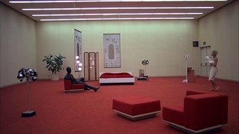 Movie still from “Death Race 2000” (1975), directed by Paul Bartel – A man sitting on a red chair in a room; Extreme Wide shot, High angle